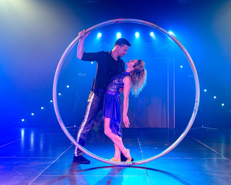 Ein Mann und eine Frau führen eine dramatische Tanzpose in einem großen Metallreifen auf einer Bühne mit bunter blauer und lila Beleuchtung auf. Die Frau lehnt sich anmutig zurück, während der Mann den Reif hält.