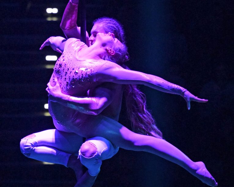 Zwei Luftakrobaten nehmen eine elegante Pose in der Luft ein, wobei die dramatische Beleuchtung ihre Kraft und Anmut vor einem dunklen Hintergrund hervorhebt.