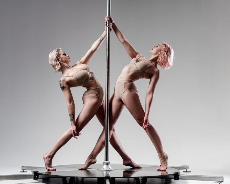 Zwei Frauen in nudefarbenen Outfits führen eine synchrone Stangentanznummer auf, wobei sie sich nach hinten lehnen und sich an einer vertikalen Stange auf einer runden Plattform vor einem hellgrauen Hintergrund festhalten.