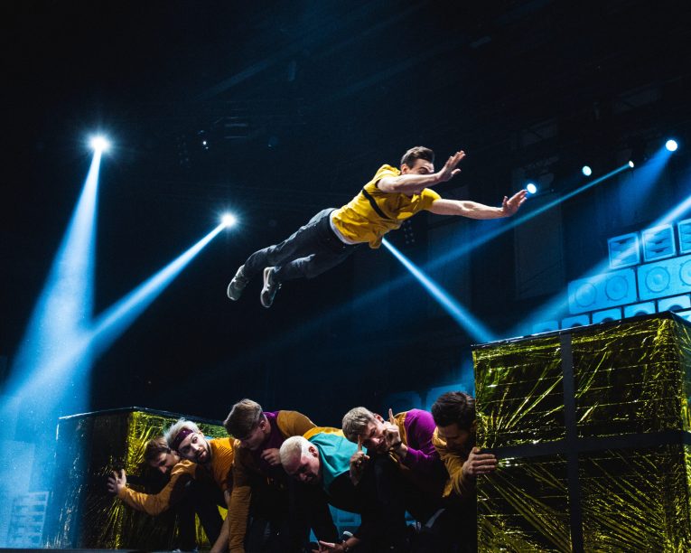 Ein Darsteller in einem gelben Hemd springt dramatisch über eine Gruppe von Menschen, die auf der Bühne zusammengekauert sind, während im Hintergrund helle Scheinwerfer leuchten und große goldbedeckte Kisten zu sehen sind.