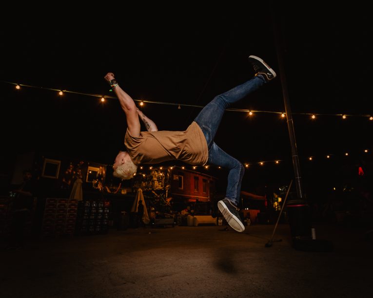 Eine Person mit gebleichtem Haar, die ein braunes T-Shirt und Jeans trägt, vollführt nachts im Freien unter Lichterketten einen akrobatischen Salto, während im Hintergrund Gebäude und Festivaldekorationen zu sehen sind.