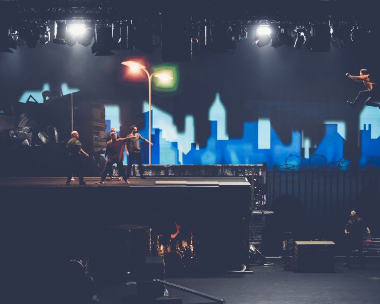 Vier Personen stehen auf einem Bühnenbild, das an ein nächtliches Stadtdach erinnert, während eine Person dramatisch durch die Luft springt. Im Hintergrund sind eine Stadtsilhouette und Straßenlaternen zu sehen, über der Bühne sind Scheinwerfer angebracht.