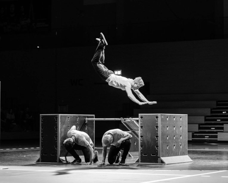 Eine Person vollführt in der Luft einen Salto über zwei kauernde Personen, die sich alle auf dem Boden einer Turnhalle befinden, die von großen, kastenförmigen Hindernissen umgeben ist. Die Szene ist in Schwarz-Weiß gehalten, im Hintergrund ist ein Publikum zu sehen.