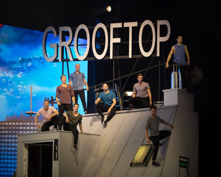 Acht Männer posieren dynamisch auf einem schrägen Dach, über dem ein großes Schild mit der Aufschrift CROOFTOP angebracht ist. Im Hintergrund sind städtische Gebäude und ein blauer Himmel zu sehen, was eine urbane Performance-Atmosphäre für die Tricking Show erzeugt.