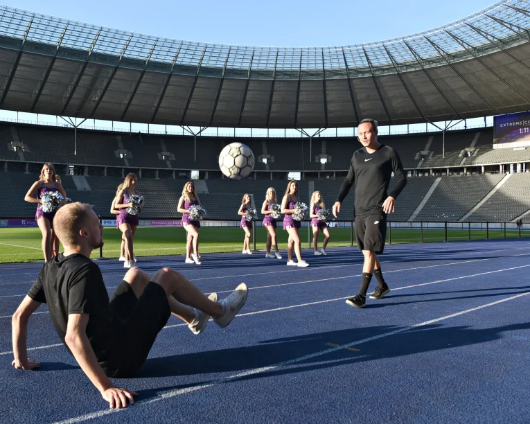 Ein Fußball-Freestyler sitzt auf einer Stadionbahn und führt gekonnt einen Fußballtrick aus, während ein anderer in der Nähe steht. Ein großer Fußball schwebt in der Luft, während Cheerleader in lila Outfits im Hintergrund des leeren Stadions stehen.