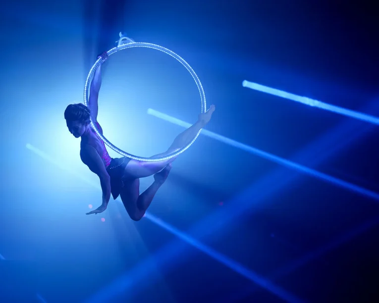 Eine Luftakrobatin posiert anmutig in einem schwebenden LED-Aerial Hoop, der von dramatischen blauen Bühnenlichtern und Strahlen beleuchtet wird, was eine eindrucksvolle und künstlerische Szene ergibt.