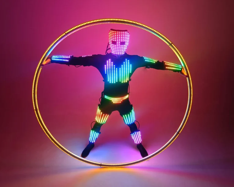 Eine Person in einem Anzug, der mit bunten LED-Lichtern bedeckt ist, steht mit ausgestreckten Armen in einem großen beleuchteten LED-Cyr-Rad vor einem Hintergrund mit Farbverlauf in Pink und Orange.