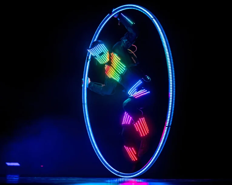 Ein Darsteller in einem Anzug mit mehrfarbigen LED-Lichtern dreht sich in einem großen, leuchtenden LED-Cyr-Rad vor einem dunklen Hintergrund und erzeugt so einen lebendigen, futuristischen visuellen Effekt.