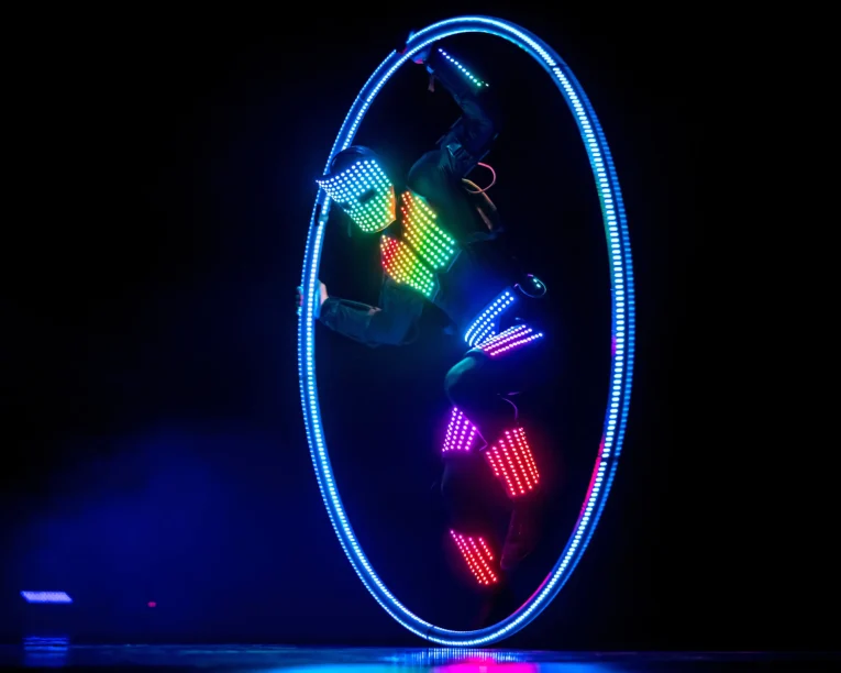 Ein Darsteller in einem Anzug mit mehrfarbigen LED-Lichtern dreht sich in einem großen, leuchtenden LED-Cyr-Rad vor einem dunklen Hintergrund und erzeugt so einen lebendigen, futuristischen visuellen Effekt.