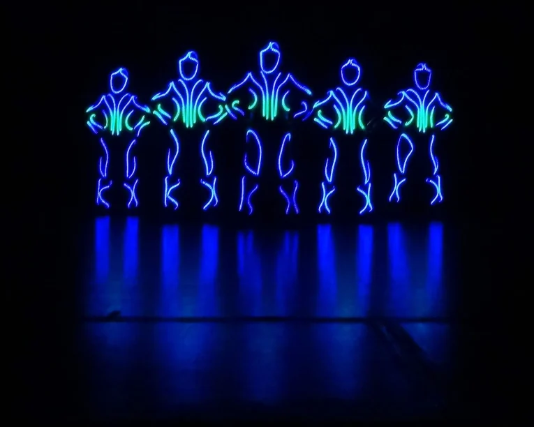 Fünf menschliche Figuren, die mit blauen und grünen LED-Lichtern umrandet sind, stehen in der Dunkelheit nebeneinander und bilden leuchtende Silhouetten mit den Händen in den Hüften, die sich auf dem glänzenden Boden spiegeln - die Essenz eines LED-Showtanzes.