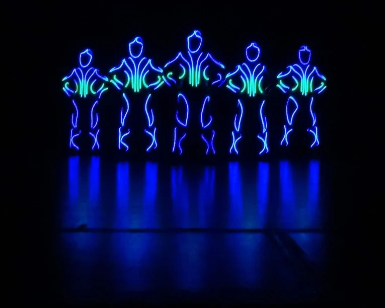 Fünf menschliche Figuren, die mit blauen und grünen LED-Lichtern umrandet sind, stehen in der Dunkelheit nebeneinander und bilden leuchtende Silhouetten mit den Händen in den Hüften, die sich auf dem glänzenden Boden spiegeln - die Essenz eines LED-Showtanzes.