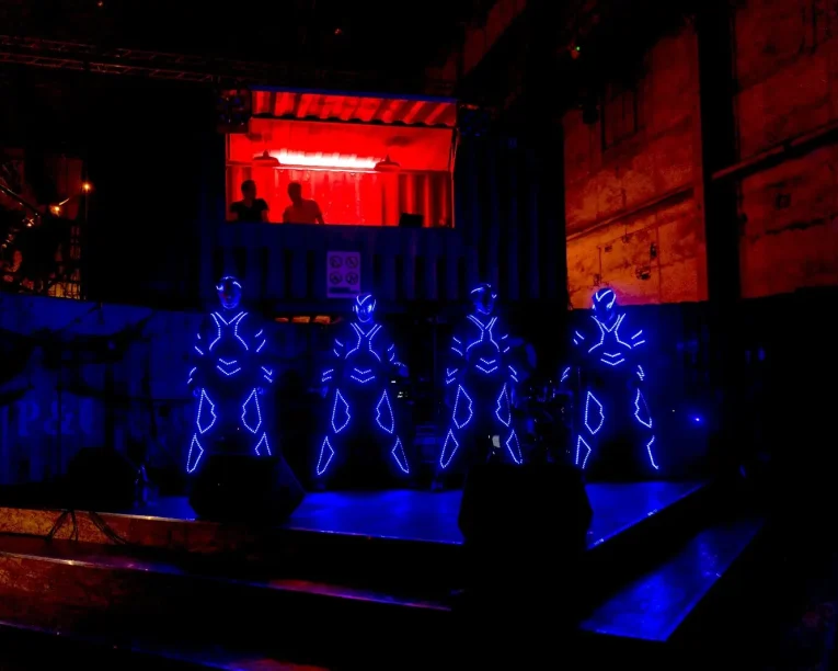 Vier Performer in leuchtenden, futuristischen Anzügen mit blauen LED-Lichtern führen auf einer schwach beleuchteten Bühne einen hypnotisierenden LED-SHOW-TANZ auf, während die Zuschauer von einem rot beleuchteten Balkon in einem scheinbar industriellen oder lagerähnlichen Raum zusehen.