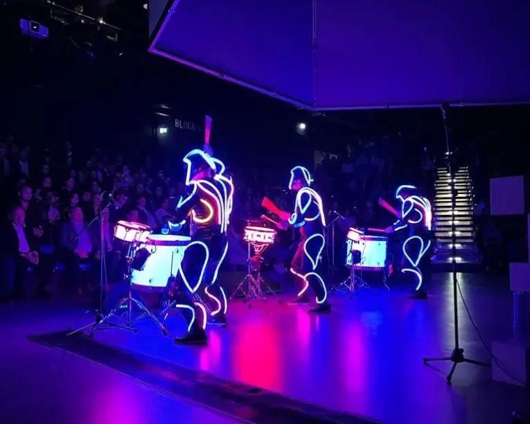 Drei Performer in leuchtenden, neonbeleuchteten Anzügen liefern eine elektrisierende LED-Trommelshow auf einer dunklen Bühne, die ein leuchtendes blaues und rosa Licht ausstrahlt. Das Publikum schaut von links zu, während im Hintergrund helle Stufen beleuchtet werden.