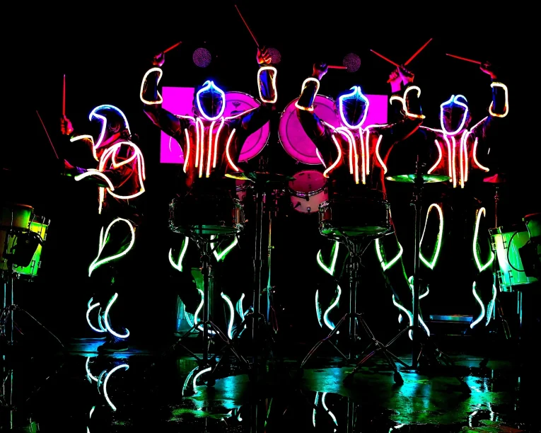 Vier Schlagzeuger treten auf der Bühne im Dunkeln auf und tragen leuchtende Neonanzüge in Rot, Blau und Grün. Mit leuchtenden Schlagzeugstöcken erzeugt diese LED-Trommel-Show einen auffälligen, futuristischen Effekt vor dem schwarzen Hintergrund.