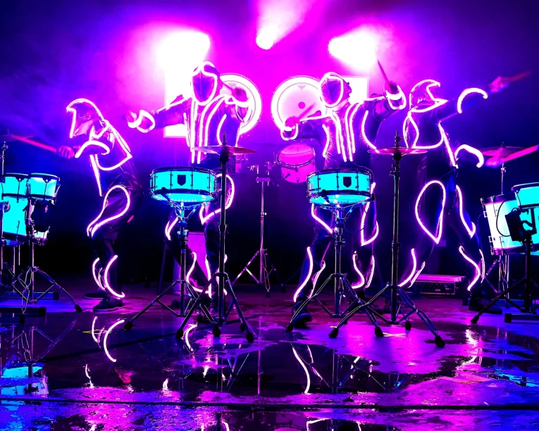 Fünf Schlagzeuger in leuchtenden Neonanzügen führen eine elektrisierende LED-Trommelshow auf der Bühne mit hellen lila und pinken Lichtern auf, umgeben von Schlagzeugsets. Der glänzende Boden spiegelt die leuchtenden Farben und die dynamische Energie der Szene wider.