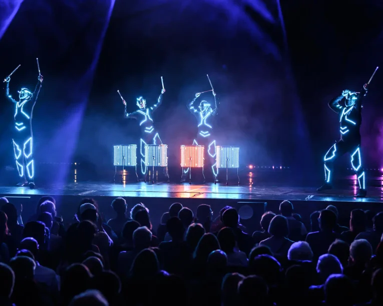 Drei Darsteller in leuchtenden, futuristischen Anzügen spielen auf einer dunklen Bühne vor einem begeisterten Publikum auf beleuchteten Trommeln, während leuchtende Lichter den Rahmen für diese elektrisierende LED Drum & Dance Show bilden.