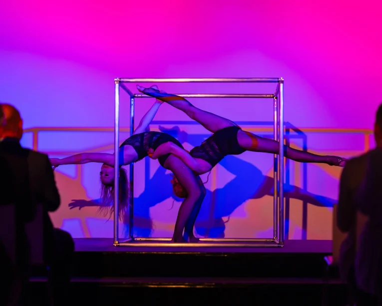 Zwei Akrobaten in schwarzen Kostümen zeigen eine Duo Acrobatic Cube Show, bei der sie in einem Metallwürfel balancieren und ihre Körper in entgegengesetzte Richtungen strecken. Die Bühne erstrahlt in leuchtendem Pink und Violett, während die Silhouetten des Publikums zusehen.