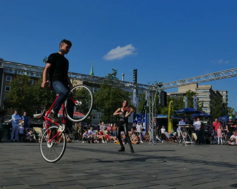 Ein Straßenkünstler verblüfft die Menge bei der Violine & Fahrrad Show, indem er auf dem Vorderrad eines Fahrrads balanciert, während eine andere Person zusieht. Im Hintergrund genießen die Menschen die sonnige Freiluftveranstaltung unter einem strahlend blauen Himmel.