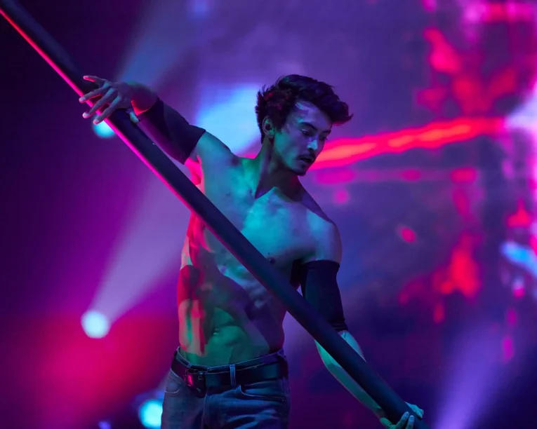 Ein hemdsärmeliger männlicher Darsteller hält sich während einer Flying Pole Show auf der Bühne an einer Stange fest, die von leuchtenden violetten, blauen und rosafarbenen Lichtern beleuchtet wird, mit einem dramatischen und intensiven Hintergrund.
