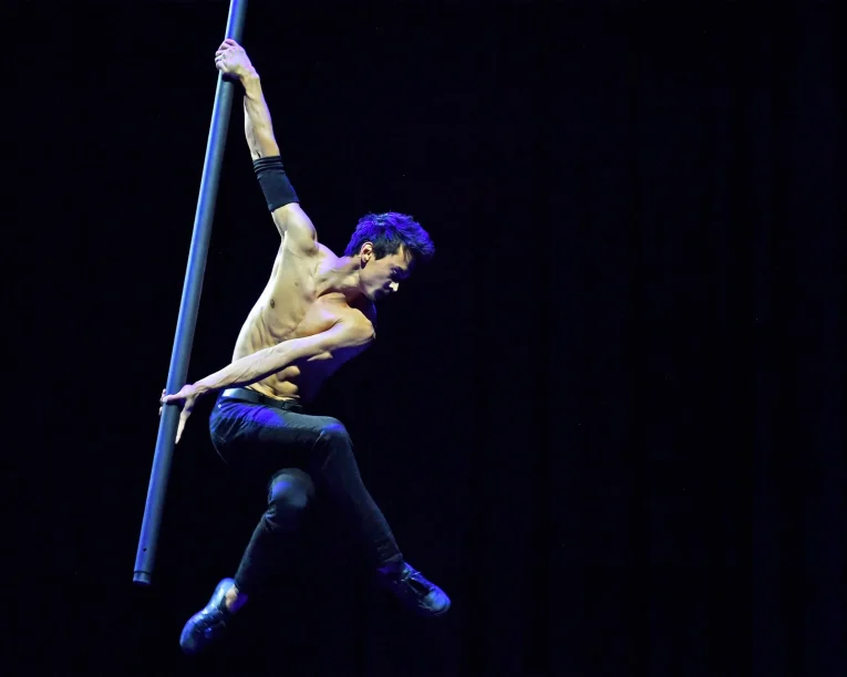 Ein hemdsärmeliger Akrobat in schwarzen Hosen und Schuhen führt vor einem dunklen Hintergrund eine Flying Pole Show vor, wobei er die vertikale Stange mit einer Hand festhält und die Beine in der Luft ausstreckt.