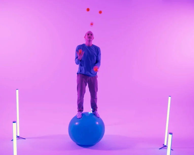 Eine Person führt eine Gymnastikball-Show vor, bei der sie auf einem großen blauen Ball steht und mit orangefarbenen Bällen in einem mit lila und rosa Lichtern beleuchteten Raum jongliert, mit zwei vertikalen Lichtbalken auf beiden Seiten.