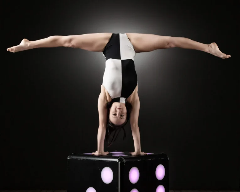 Eine Frau in einem schwarz-weißen Trikot zeigt atemberaubende Handstandakrobatik, indem sie einen breitbeinigen Handstand auf einem großen, beleuchteten Würfel auf einem Holzboden vor einem dunklen Hintergrund vorführt.