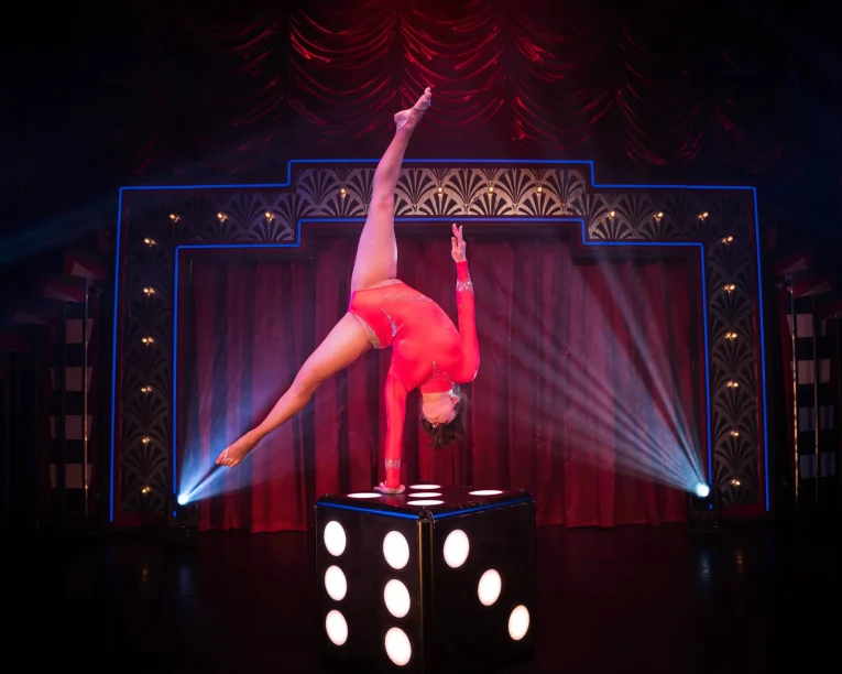 Eine Darstellerin in einem roten Trikot zeigt Handstandakrobatik, indem sie auf einer Hand auf einem großen schwarzen Würfel mit weißen Punkten balanciert, umrahmt von dramatischer Bühnenbeleuchtung und einem roten Vorhang im Hintergrund.