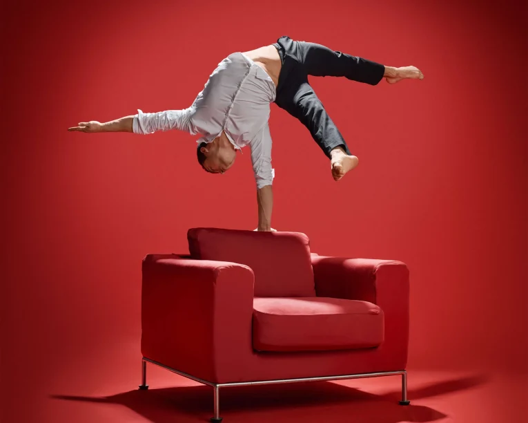 Ein Mann in weißem Hemd und schwarzer Hose führt Handstandakrobatik vor, wobei er auf einer Hand auf einem roten Sessel balanciert und einen beeindruckenden akrobatischen Handstand vor einem leuchtend roten Hintergrund vollführt.