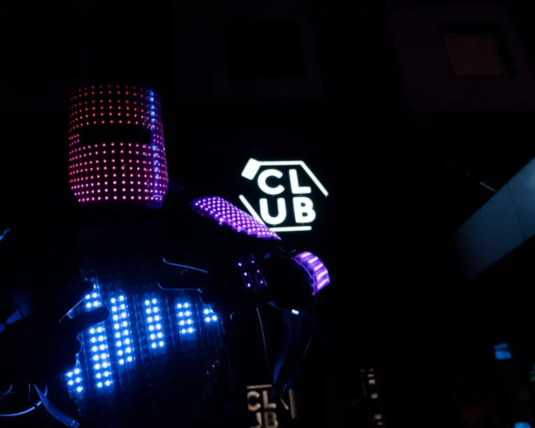 Eine Person in einem futuristischen LED-Roboter-Kostüm, das mit bunten Lichtern bedeckt ist, steht vor einem Gebäude mit einem leuchtenden CLUB-Schild und schafft eine lebendige und hochtechnologische Atmosphäre bei Nacht.
