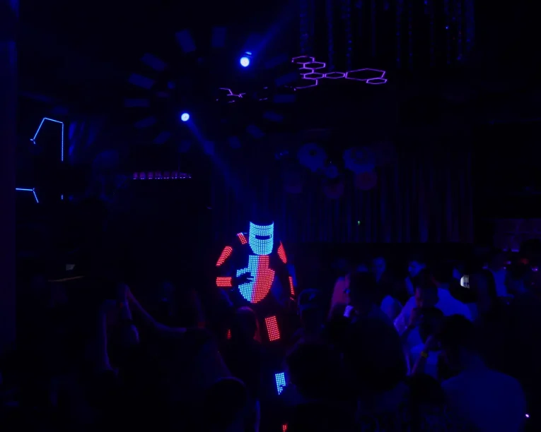 Ein LED-Roboter in einem leuchtenden Anzug tanzt in einem dunklen Nachtclub, der von blauen und roten Lichtern erhellt wird, während eine Menschenmenge zusieht und bunte Lichter die Decke schmücken.