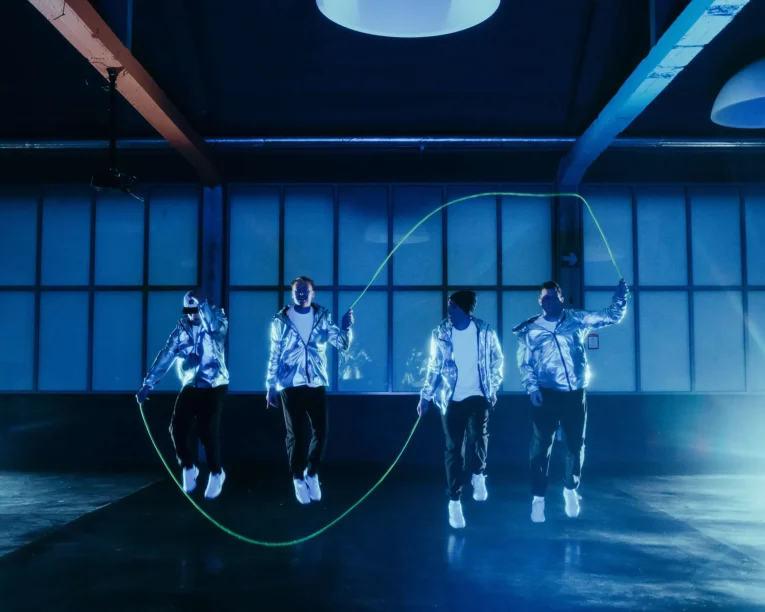 Vier Personen in glänzenden silbernen Jacken springen in einem dunklen Raum bei blauer Beleuchtung gemeinsam Seil, während sie ein großes grünes Springseil springen und drehen.