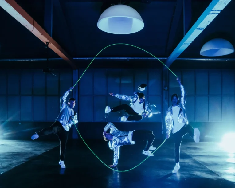 Vier Tänzerinnen und Tänzer in glänzenden silbernen Jacken führen in einem dunklen, blau beleuchteten Raum eine energiegeladene Seilsprungnummer auf, wobei zwei das Seil halten, während die anderen springen und in der Luft posieren. Helle Lichter erhellen den Hintergrund.