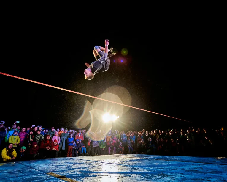 Eine Person vollführt nachts einen Rückwärtssalto auf einer Slackline, die über einer Menge von Zuschauern hängt. Helle Lichter und Streulicht beleuchten die Szene und heben die Bewegung des Slackliners in der Luft hervor.