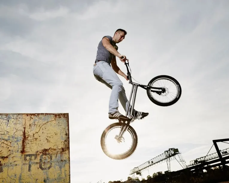 Eine Person führt während einer Trial Bike Show auf einem BMX-Rad in der Nähe einer mit Graffiti bedeckten Rampe einen Hochsprungtrick aus, wobei sich die Silhouette gegen den Himmel abhebt und im Hintergrund Industrieanlagen zu sehen sind.