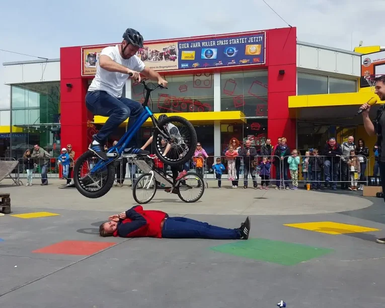 Während einer aufregenden Trial Bike Show springt ein Mann auf einem BMX-Rad über eine am Boden liegende Person, während eine Menschenmenge hinter einer Absperrung zuschaut und ein buntes Gebäude eine lebendige Kulisse bildet. Ein anderer Mann steht in der Nähe und hält ein Mikrofon.