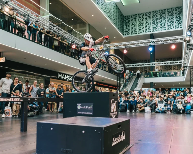 Ein Radfahrer, der einen Helm trägt, vollführt während einer Trial Bike Show einen waghalsigen Stunt. Er balanciert auf einem Rad auf einer erhöhten Plattform in einem Einkaufszentrum, während eine große Menschenmenge zusieht und Fotos macht.