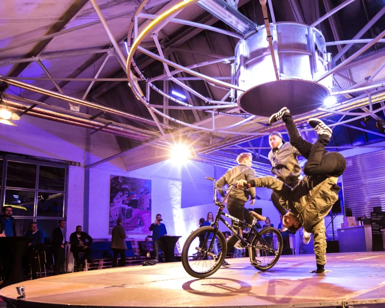 Zwei BMX-Fahrer und ein Breakdancer führen in einer Halle unter Industriebeleuchtung Stunts auf. Ein Fahrer macht einen Handstand auf dem Boden, während der andere auf einem Fahrrad steht. Die Zuschauer sehen zu, wie sich die lebhafte, farbenfrohe Atmosphäre entfaltet.