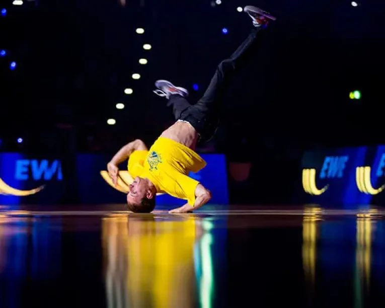 Ein Breakdancer in einem gelben Hemd vollführt einen einhändigen Headspin auf einer glänzenden Bühne und bringt energiegeladene Unterhaltung ins Rampenlicht, mit hellen Lichtern und einem dunklen Publikum im Hintergrund.