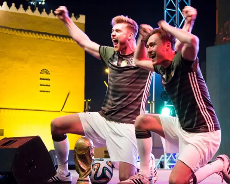 Zwei aufgeregte Football-Freestyle-Fußballer in passenden Uniformen knien auf der Bühne und heben jubelnd die Arme neben einer Nachbildung der FIFA-Weltmeisterschaftstrophäe, während hinter ihnen ein gelb beleuchtetes Gebäude die Nacht erhellt.