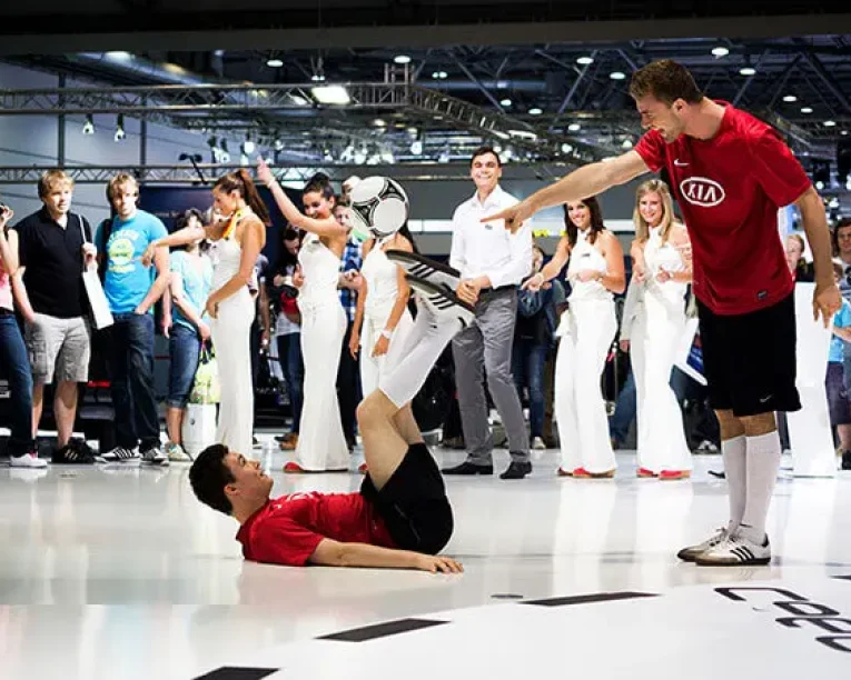 Zwei Fußballartisten in roten Trikots führen bei einem Hallenturnier eine Fußballkür vor; der eine balanciert einen Ball auf den Füßen, während der andere im Liegen auf den Ball zeigt, während die Zuschauer im Hintergrund zusehen.