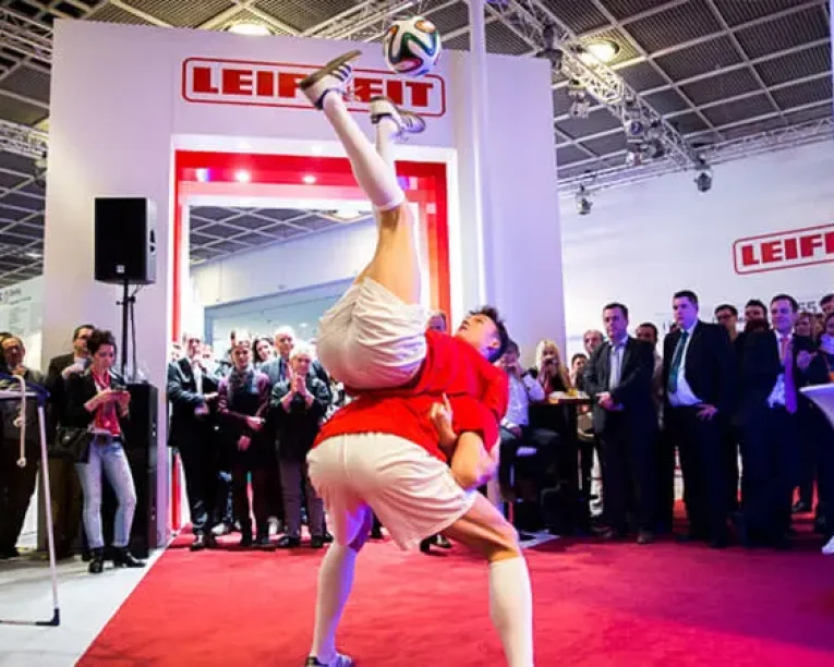 Zwei Fußballer in rot-weißen Trikots beeindrucken das Publikum bei einer Hallenveranstaltung mit einer akrobatischen Fußball-Show - einer balanciert kopfüber auf dem Rücken des anderen und hält einen Fußball gekonnt mit den Füßen fest.