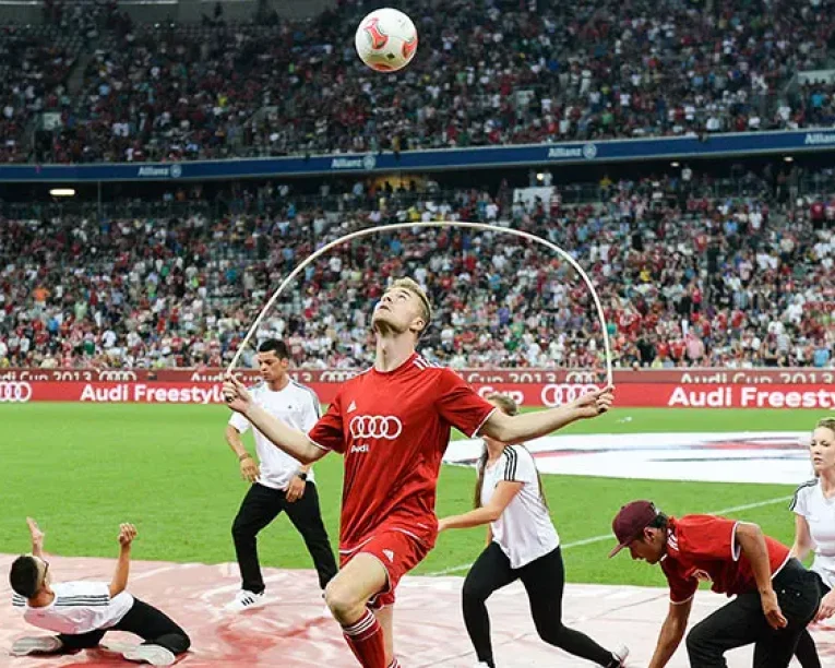 Ein Mann in einem roten Audi-Trikot balanciert einen Fußball auf dem Kopf, während er Seil springt und Urban-Dance-Bewegungen inmitten von Künstlern auf einem Stadionfeld macht, während eine große jubelnde Menge zusieht.