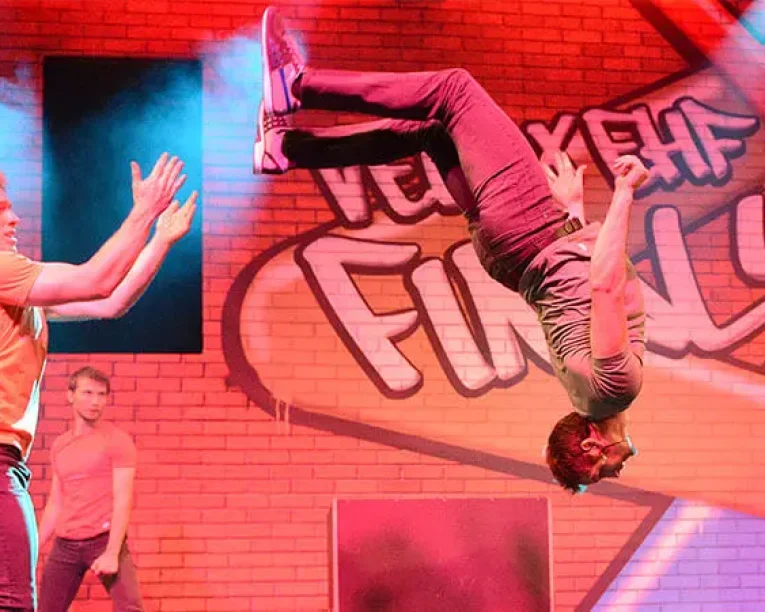 Ein Mann vollführt während einer Parkour-Show einen Rückwärtssalto auf der Bühne, während ein anderer Mann in der Nähe klatscht. Eine dritte Person steht im Hintergrund. Bunte Beleuchtung und eine Backsteinmauer mit fetten Graffiti-Schriftzügen schaffen eine lebendige Atmosphäre.