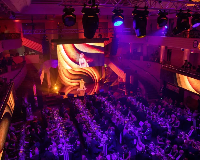 Ein großer, eleganter Bankettsaal, gefüllt mit sitzenden Gästen an langen Tischen, beleuchtet von lila und goldenen Lichtern. Ein Darsteller betritt die Bühne für die McDonalds CIRCLE SHOW, während wirbelnde goldene Muster über den großen Bildschirm hinter ihm tanzen.