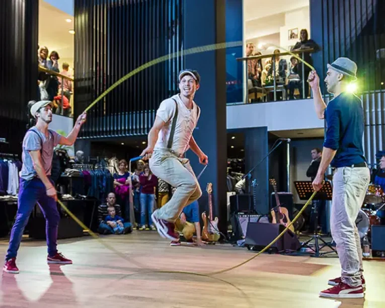 Drei Männer mit Hüten führen einen doppelten holländischen Seilsprung in der Halle durch und stellen ihr Supertalent unter Beweis, indem einer springt, während die anderen die Seile drehen. Die Zuschauer schauen vom Hintergrund und vom oberen Balkon aus zu, während rechts eine Musikkapelle spielt.