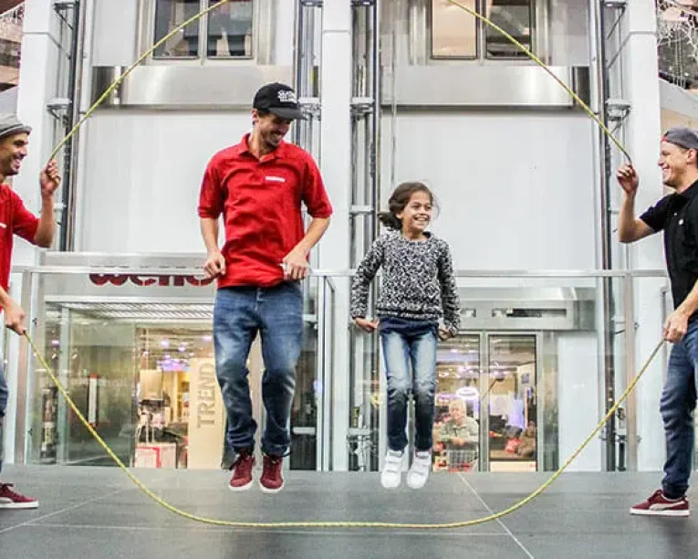 Drei Männer in roten Hemden und Hüten schwingen ein großes Springseil, während ein lächelnder junger Ropeskipper in Pullover und Jeans in der Mitte springt, alles auf einer Bühne in einem modernen Einkaufszentrum.