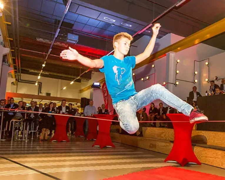 Ein junger Mann in blauem T-Shirt und Jeans springt während einer Indoor-Slackline-Show in der Luft über eine Slackline, während die Zuschauer von erhöhten Sitzen und einem Barbereich aus zusehen. Der hell erleuchtete Raum wird von Scheinwerfern erhellt.