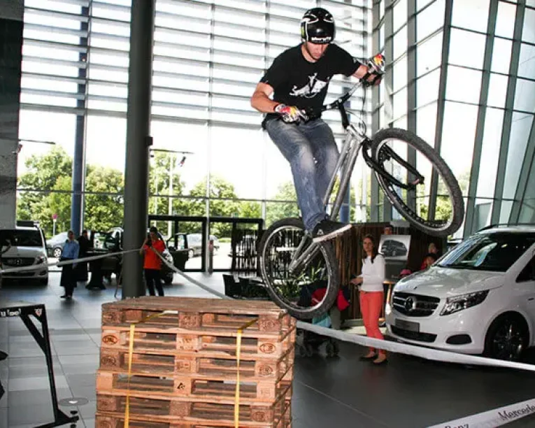 Ein Radfahrer mit Helm vollführt einen aufregenden Stunt auf einem Trial-Bike, indem er über gestapelte Holzpaletten in einer Halle springt, während im Hintergrund Autos und Menschen zusehen.