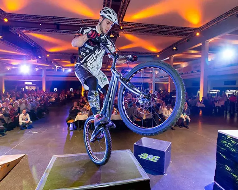 Ein Radfahrer in voller Montur vollführt auf der Bühne der Fahrradmesse einen Wheelie-Stunt, bei dem er das Vorderrad des Fahrrads anhebt, während ein großes Publikum unter bunten Lichtern in einer Halle zusieht.