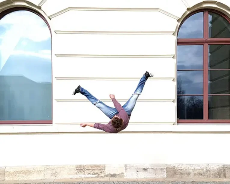 Eine Person, die Free Running praktiziert, scheint seitlich auf einer weißen Gebäudewand zwischen zwei großen Bogenfenstern zu liegen, wobei ihre Beine in der Luft gespreizt sind, was eine optische Täuschung erzeugt, als ob die Schwerkraft seitwärts wirkt.